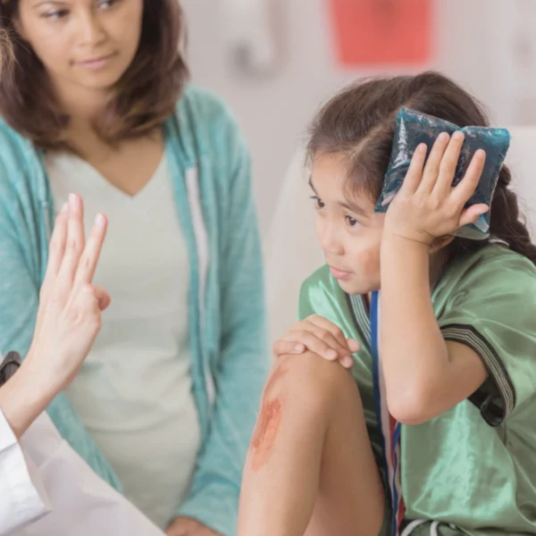 Enfant blessée après un accident de la route, examinée par un médecin avec des éraflures visibles et une poche froide sur le front