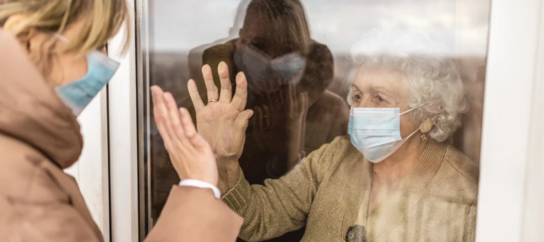 Deux proches séparés par une vitre avec masque, illustrant l’isolement des victimes du Covid-19 et le préjudice moral des ayants droit des victimes décédées