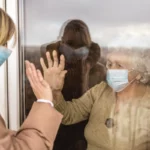 Deux proches séparés par une vitre avec masque, illustrant l’isolement des victimes du Covid-19 et le préjudice moral des ayants droit des victimes décédées